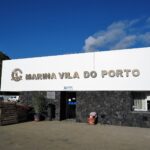 The reception building of the Vila do Porto marina, a large white sign with metal letters on a black basalt wall, under a blue sky, with a green cliff on the left