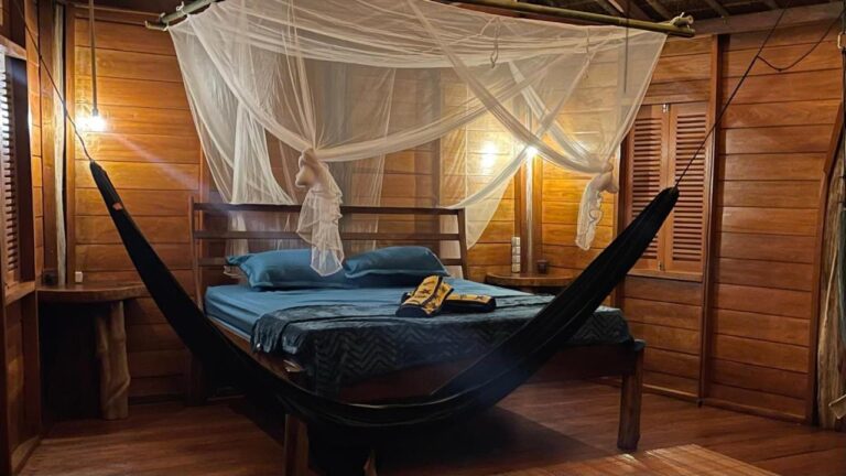 A room with wooden walls lit by lamps, with a bed under a mosquito net and a black hammock