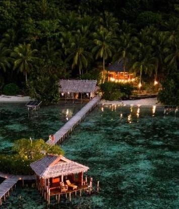 Aerial view of Nyande Raja Ampat at dusk, with a stilt bungalow and the jetty leading to the beach