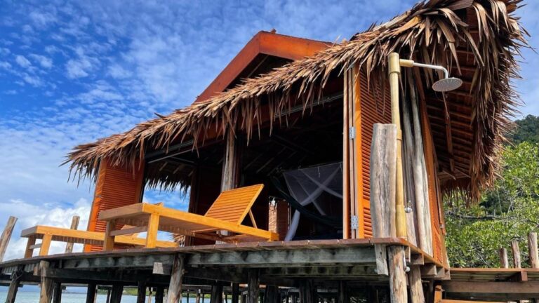 A stilt bungalow with a palm-thatched roof, two wooden sun loungers and an outdoor shower