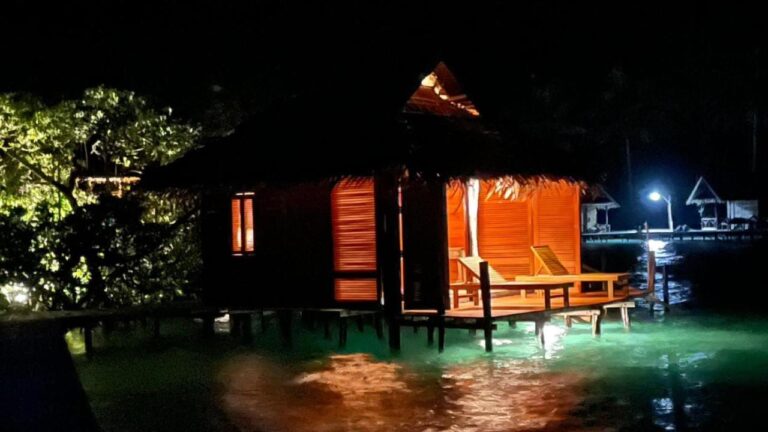 A stilt bungalow lit up at night, with sun loungers on its terrace