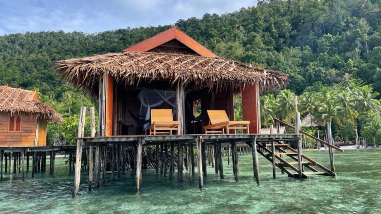 A stilt bungalow with a palm-thatched roof, two sun loungers and a staircase down into the water