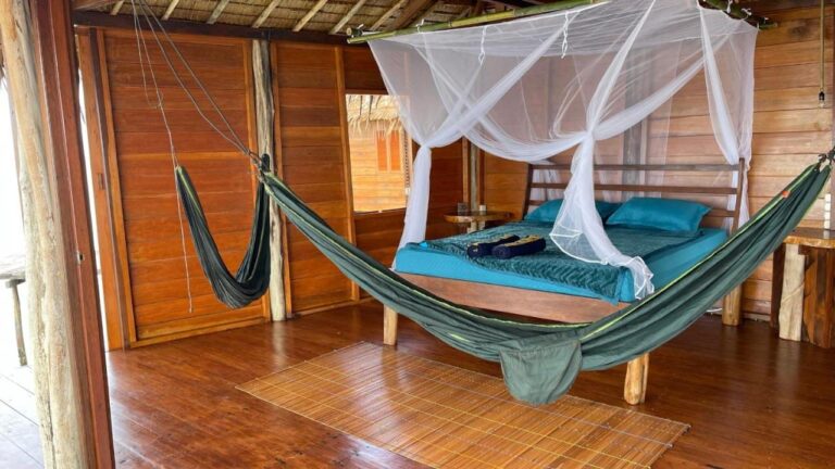 A wooden room open to the outside, with a bed with blue sheets under a mosquito net and a green hammock