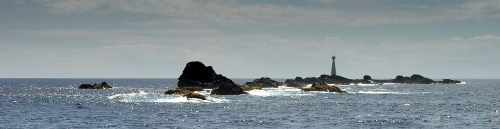 Panorama of the chain of black Formigas reefs at water level, edged with white foam on a sparkling blue sea, with the white Formigão lighthouse on the right under a hazy sky