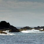 Panorama of the chain of black Formigas reefs at water level, edged with white foam on a sparkling blue sea, with the white Formigão lighthouse on the right under a hazy sky