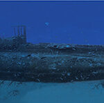 Underwater panorama of the full length of the Rubis wreck, sitting upright on the sand, conning tower in the center, in blue water dotted with fish.