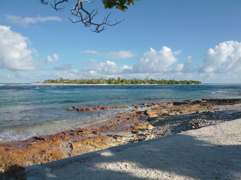 The pass channel with choppy water, between a reddish rock flat in the foreground and a low islet covered in coconut palms on the far shore, under a sky of cumulus clouds