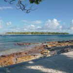 The pass channel with choppy water, between a reddish rock flat in the foreground and a low islet covered in coconut palms on the far shore, under a sky of cumulus clouds