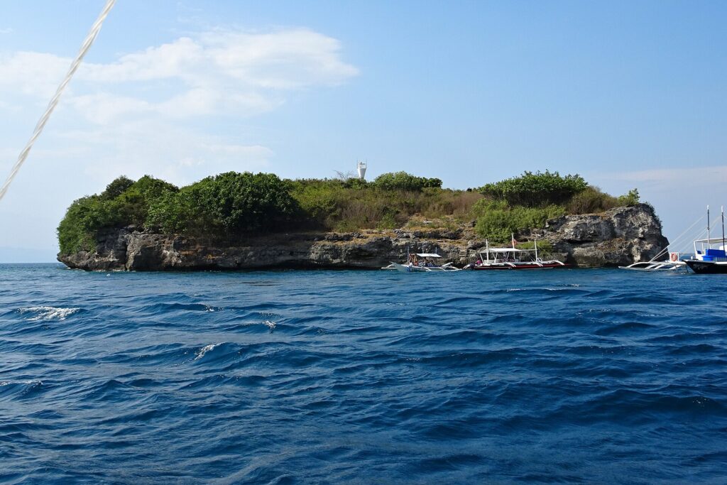 Pescador Island seen from the sea, covered in green bushes, its white lighthouse on top and three dive bancas moored at the foot of the cliff
