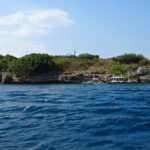 Pescador Island seen from the sea, covered in green bushes, its white lighthouse on top and three dive bancas moored at the foot of the cliff