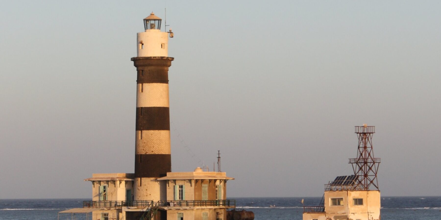 The Daedalus lighthouse, a black and white striped tower standing on the reef, with its buildings and metal jetty, at sunset