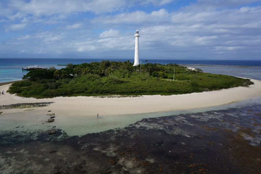 The tall white lighthouse of Amédée islet rises above the vegetation, the islet ringed with white sand, the exposed coral flat in the foreground and a boat docked on the left