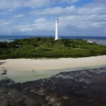 The tall white lighthouse of Amédée islet rises above the vegetation, the islet ringed with white sand, the exposed coral flat in the foreground and a boat docked on the left