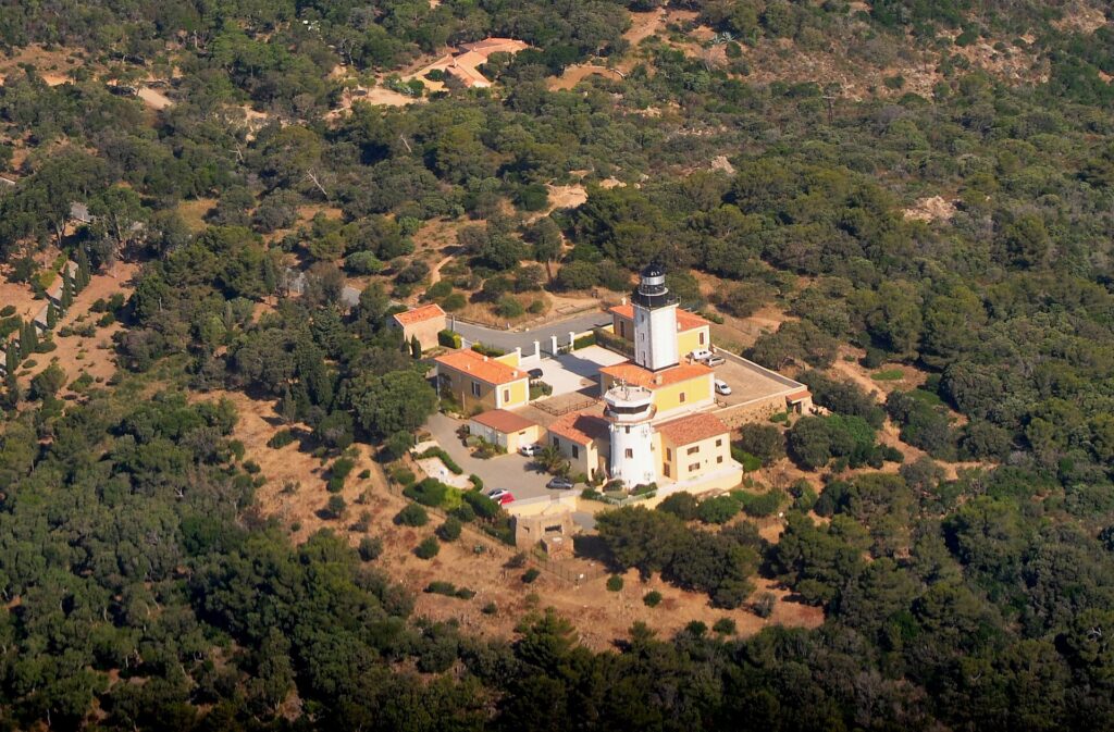 Aerial view of the Cap Camarat lighthouse, a white tower with a black lantern surrounded by yellow buildings with orange tile roofs, in the middle of a dense Mediterranean forest.