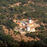 Aerial view of the Cap Camarat lighthouse, a white tower with a black lantern surrounded by yellow buildings with orange tile roofs, in the middle of a dense Mediterranean forest.