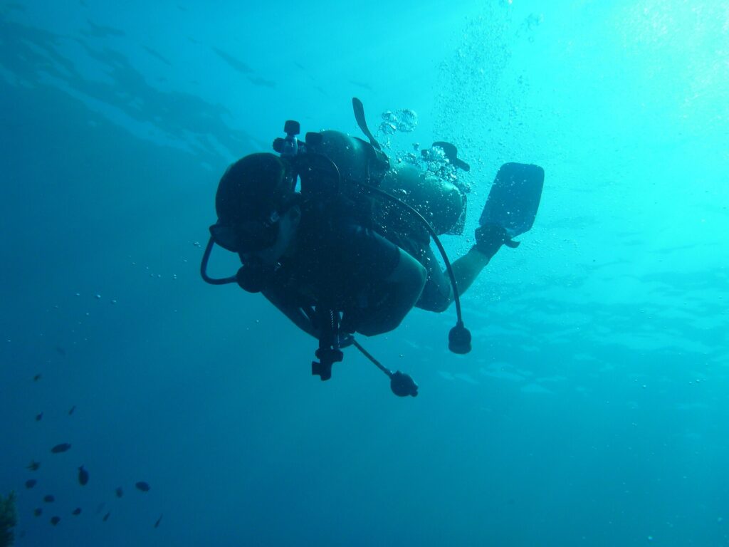 A diver in silhouette seen from below, tank, regulators and fins outlined against turquoise water, bubbles rising toward the sunlit surface.