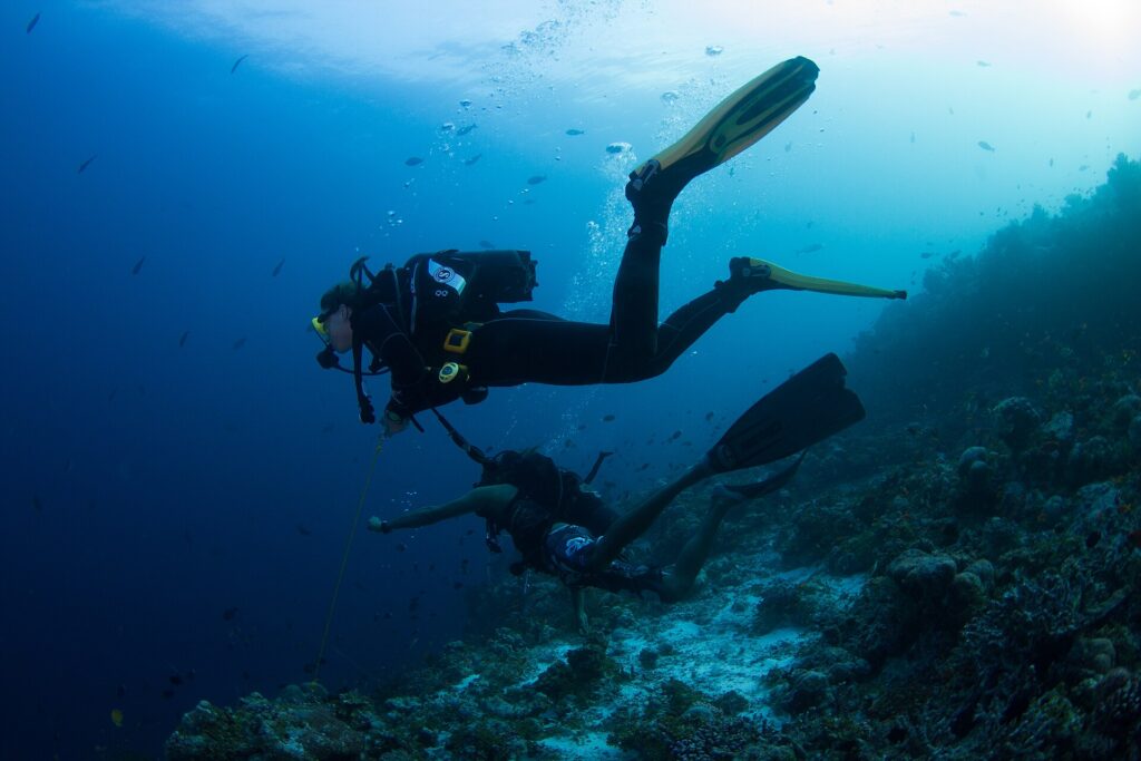 Two divers above a reef slope, one held by a reef hook line, the other in shorts, in blue water full of small fish