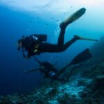 Two divers above a reef slope, one held by a reef hook line, the other in shorts, in blue water full of small fish