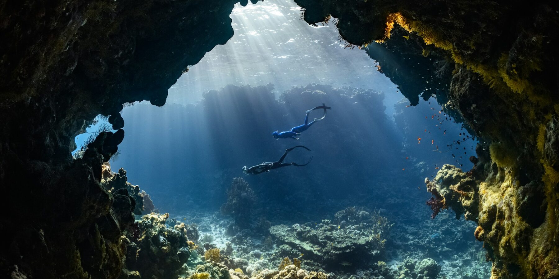 Two divers swim through an underwater cave lit by sunbeams, above a reef