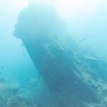 The broken stern of the Thistlegorm rising at an angle in the blue, a diver on the right