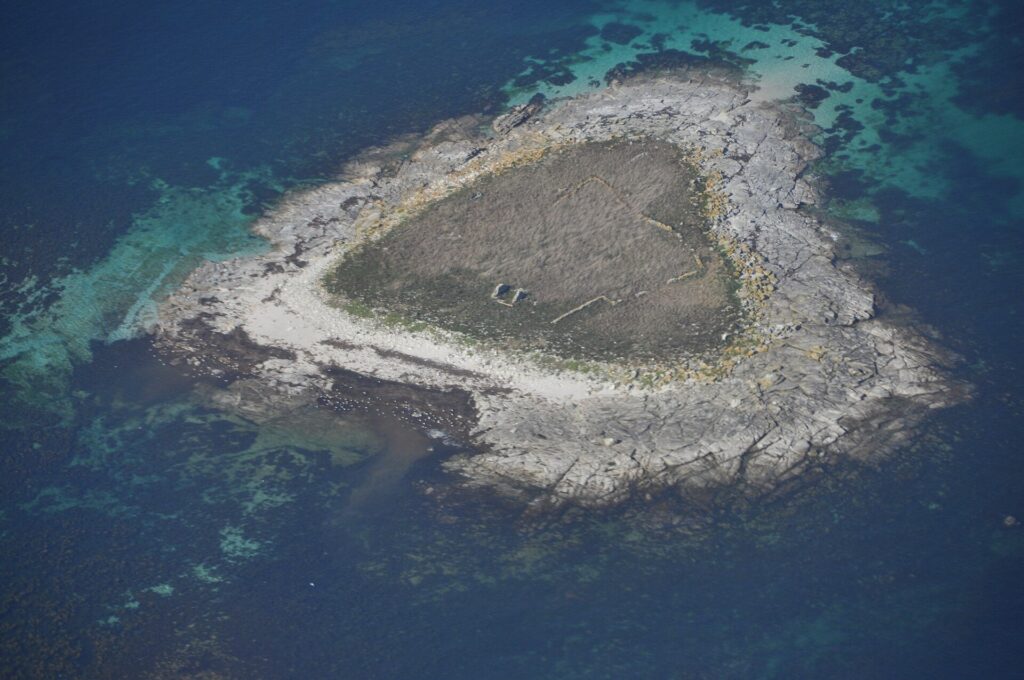 Aerial view of a small islet of gray granite slabs, covered in dry grass in the center where ruined low walls can be made out, edged by a pale beach and surrounded by turquoise shallows and deep blue water.