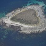 Aerial view of a small islet of gray granite slabs, covered in dry grass in the center where ruined low walls can be made out, edged by a pale beach and surrounded by turquoise shallows and deep blue water.