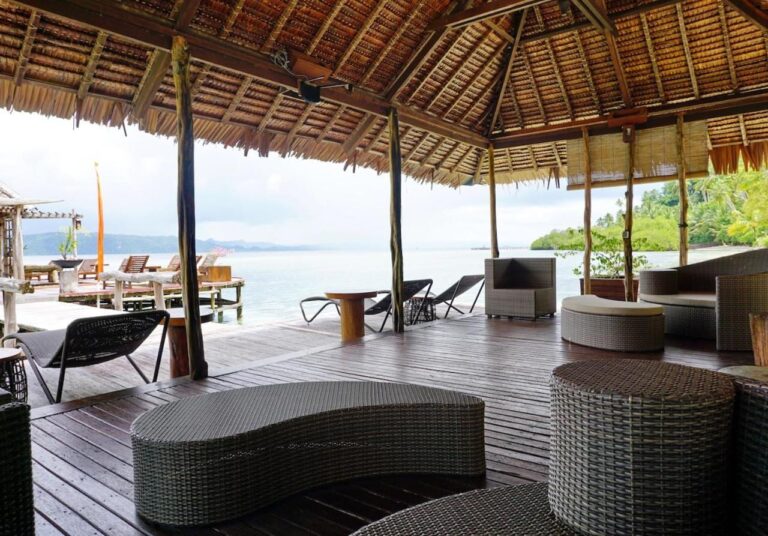 A lounge under a palm-thatched roof, with woven seats and the sea in the background