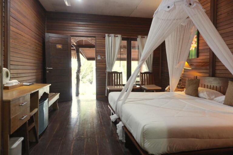 A double room with a bed under a mosquito net, a desk, a kettle and a small fridge