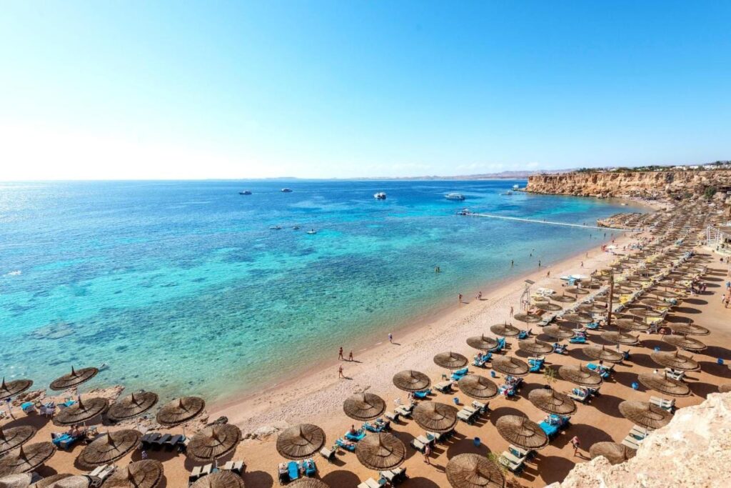 A long sandy beach at the foot of a cliff, with straw parasols and a jetty over the turquoise sea
