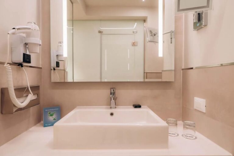 A white sink under a lit mirror, with a hairdryer on the wall