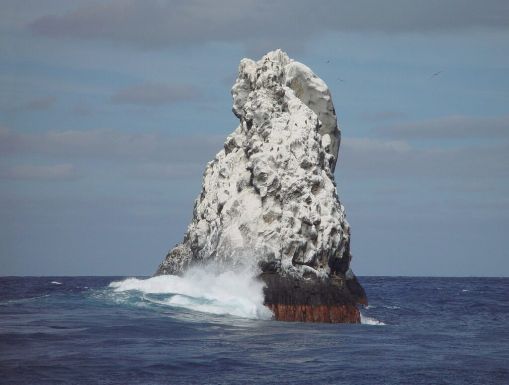The northwest peak of Roca Partida, white with guano, seen from the boat, a wave breaking at its base and birds flying above