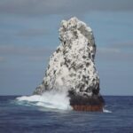 The northwest peak of Roca Partida, white with guano, seen from the boat, a wave breaking at its base and birds flying above