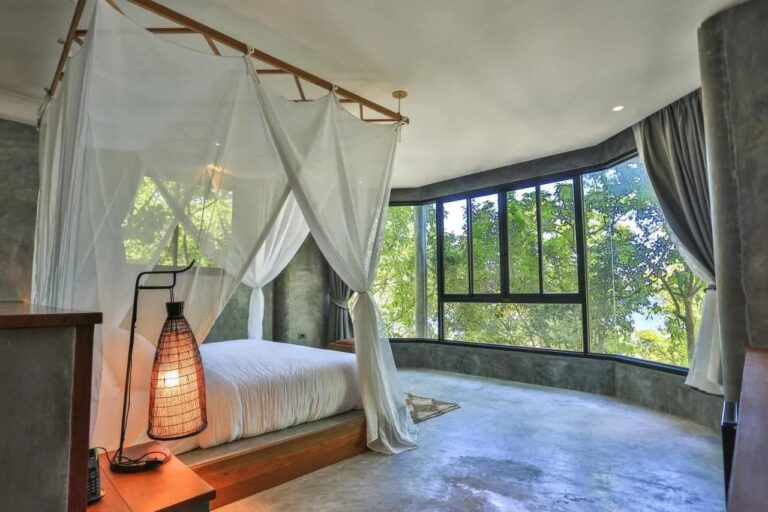A room with concrete walls, a bed under a mosquito net and a picture window onto the trees