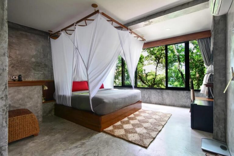 A bed under a mosquito net with red cushions, facing a large window onto the forest