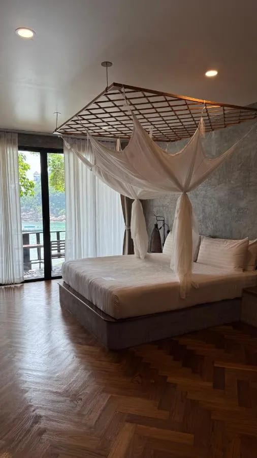 A room with a herringbone parquet floor, a bed under a mosquito net and a glass door onto the sea
