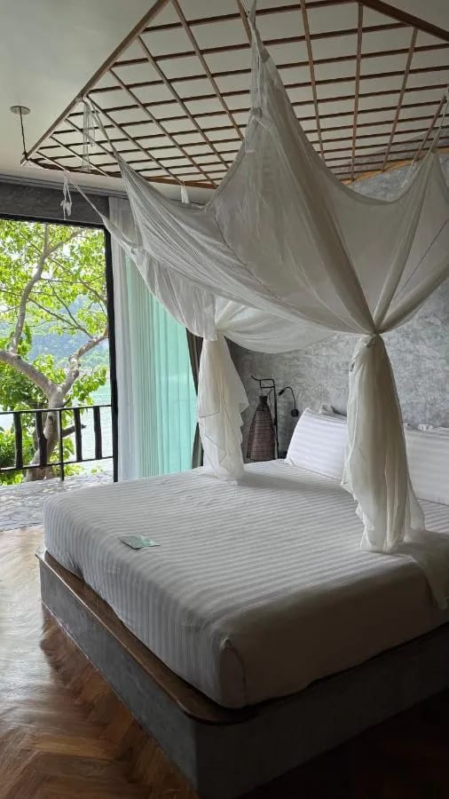 A bed under a mosquito net facing a glass door, with a balcony over the trees and the sea