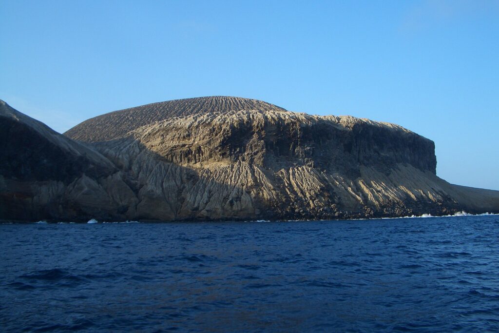 The coast of San Benedicto seen from the sea: the ash cone of Bárcena volcano, streaked with gullies, above dark cliffs and a deep blue sea
