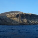 The coast of San Benedicto seen from the sea: the ash cone of Bárcena volcano, streaked with gullies, above dark cliffs and a deep blue sea