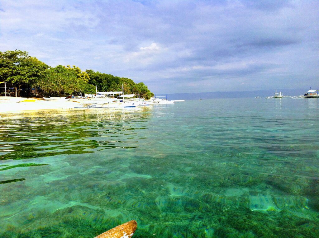 Clear turquoise water over a shallow bottom, in front of a tree-lined white sand beach where white bangkas line up, other boats offshore under a cloudy sky.