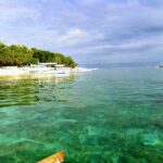 Clear turquoise water over a shallow bottom, in front of a tree-lined white sand beach where white bangkas line up, other boats offshore under a cloudy sky.
