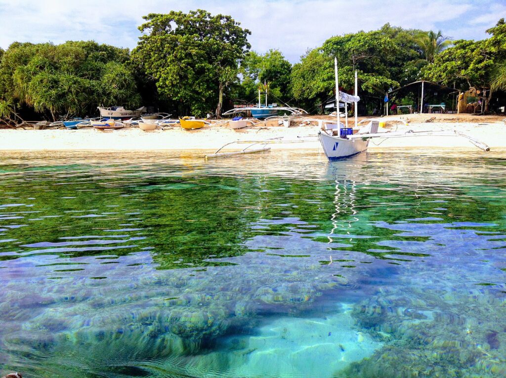 At water level, coral heads under clear turquoise water, in front of a white sand beach where a white bangka is moored, other colorful boats pulled up under the trees.