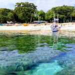 At water level, coral heads under clear turquoise water, in front of a white sand beach where a white bangka is moored, other colorful boats pulled up under the trees.