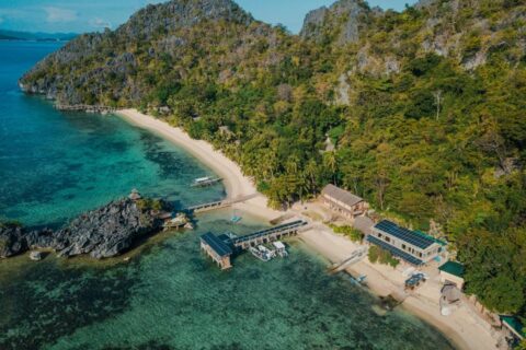 Aerial view of Sangat Island Dive Resort, with its beach, its jetty and its boats, at the foot of the cliffs