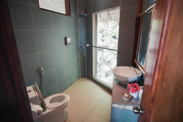 A bathroom with dark green tiles, a sink, a shower and a window onto the rocks