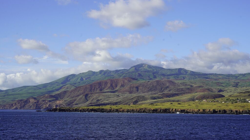 The coast of Socorro Island seen from the sea: black lava cliffs, green and reddish hills and the volcano's summit in the clouds