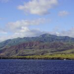 The coast of Socorro Island seen from the sea: black lava cliffs, green and reddish hills and the volcano's summit in the clouds