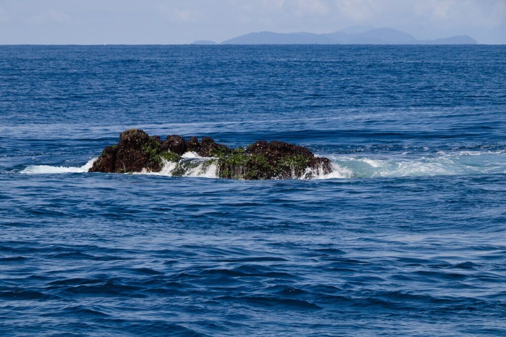 The top of Richelieu Rock breaking the surface at low tide, a dark rock covered in green algae and pounded by waves, with the Surin Islands on the horizon