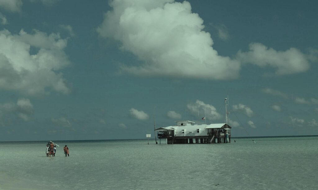 The ranger station, a white building on stilts bristling with antennas and topped with a Philippine flag, in pale green shallow water, three people in orange vests wading on the left under a big cumulus cloud.