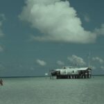The ranger station, a white building on stilts bristling with antennas and topped with a Philippine flag, in pale green shallow water, three people in orange vests wading on the left under a big cumulus cloud.