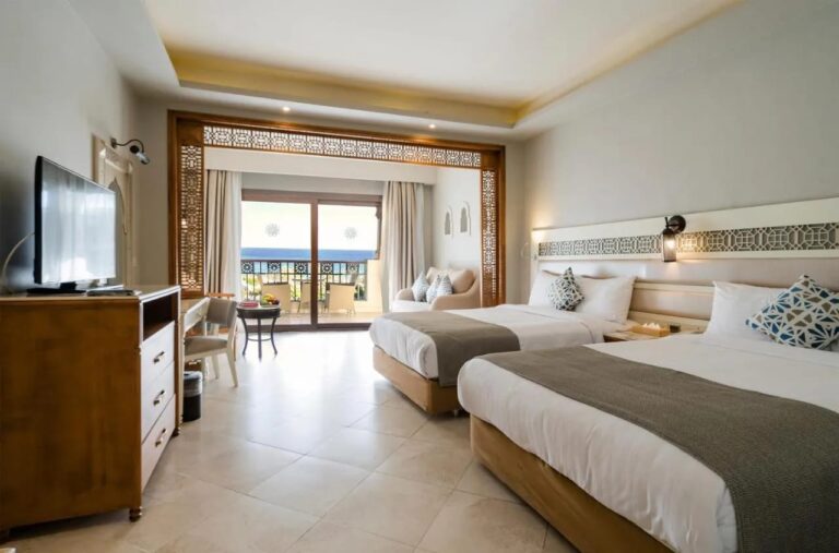 A room with two double beds, a seating area under a wooden arch and a sea-facing balcony